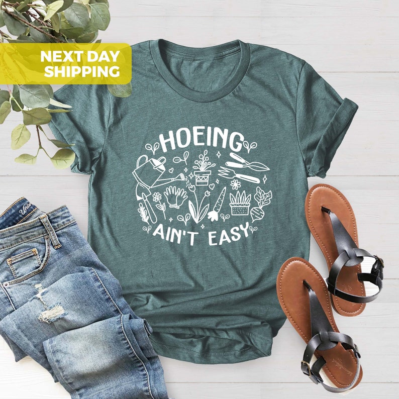 K&ouml;nnte beinhalten: Ein tealgr&uuml;nes T-Shirt mit einem wei&szlig;en Grafikdesign, auf dem "HOEING AIN'T EASY" steht, umgeben von Gartenger&auml;ten und Pflanzen.