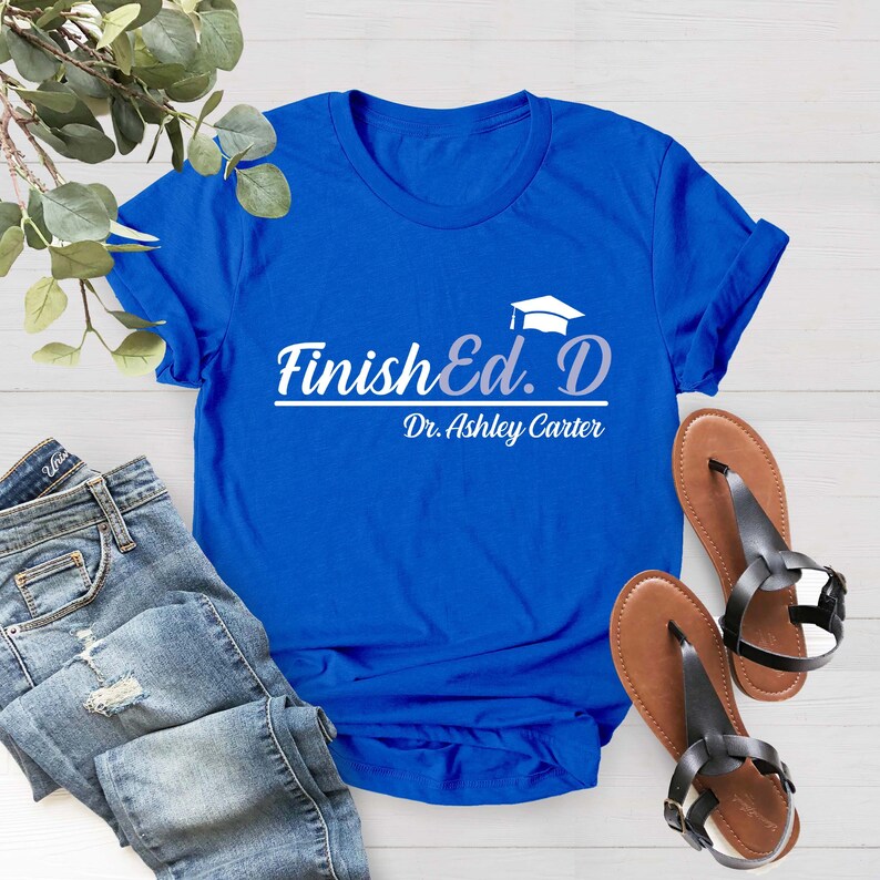 Puede incluir: Una camiseta azul real con el texto "Finish Ed. D" y "Dr. Ashley Carter" en letras blancas. El texto est&aacute; estilizado con un gr&aacute;fico de gorro de graduaci&oacute;n.