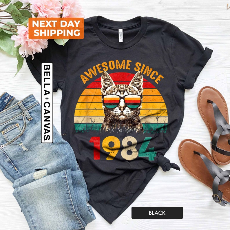Może przedstawiać: Czarny T-shirt z grafiką w stylu vintage przedstawiającą kota w okularach przeciwsłonecznych i tekstem "Awesome Since 1984".