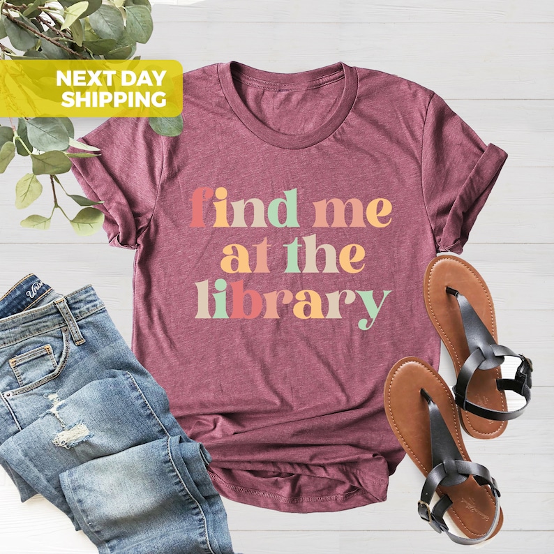 Puede incluir: Una camiseta de color granate con el texto "find me at the library" en letras de colores.