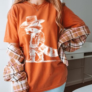 May include: A burnt orange t-shirt with a white graphic of a raccoon wearing a cowboy hat and sunglasses. The raccoon is standing with its hands on its hips.