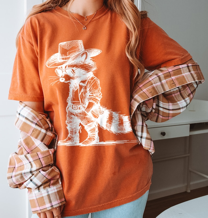May include: A burnt orange t-shirt with a white graphic of a raccoon wearing a cowboy hat and sunglasses. The raccoon is standing with its hands on its hips.