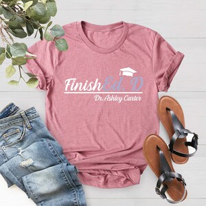 Puede incluir: Una camiseta rosa con el texto "Finished. D" y un gr&aacute;fico de un gorro de graduaci&oacute;n. El texto "Dr. Ashley Carter" est&aacute; debajo del gr&aacute;fico.