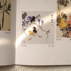 May include: A book page featuring traditional ink wash paintings. One illustration shows a yellow chick and a black bird beneath purple flowers. Another depicts a yellow gourd with pink flowers. Chinese characters and red seals are visible.