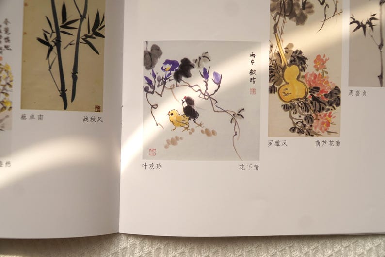 May include: A book page featuring traditional ink wash paintings. One illustration shows a yellow chick and a black bird beneath purple flowers. Another depicts a yellow gourd with pink flowers. Chinese characters and red seals are visible.