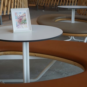 May include: A watercolour painting in a white frame sits on a small white table with a black trim. The table is supported by a white metal base. A brown circular bench surrounds the table. The table is approximately 60 cm in diameter.