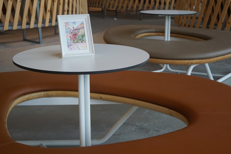 May include: A watercolour painting in a white frame sits on a small white table with a black trim. The table is supported by a white metal base. A brown circular bench surrounds the table. The table is approximately 60 cm in diameter.