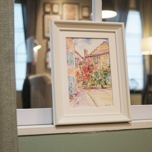 May include: A framed watercolour painting of a European street scene. The artwork depicts buildings with red-tiled roofs, blue shutters, and vibrant red flowers. The painting is in a white frame, displayed on a windowsill.