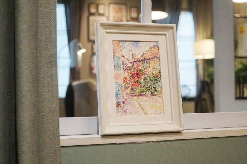 May include: A framed watercolour painting of a European street scene. The artwork depicts buildings with red-tiled roofs, blue shutters, and vibrant red flowers. The painting is in a white frame, displayed on a windowsill.