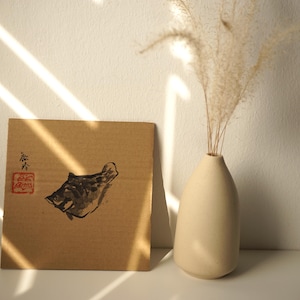 May include: A square piece of brown cardboard featuring a black ink wash painting of a fish, accompanied by a red stamp and black calligraphy. Next to it stands a smooth, matte, off-white ceramic vase containing a single sprig of dried pampas grass.