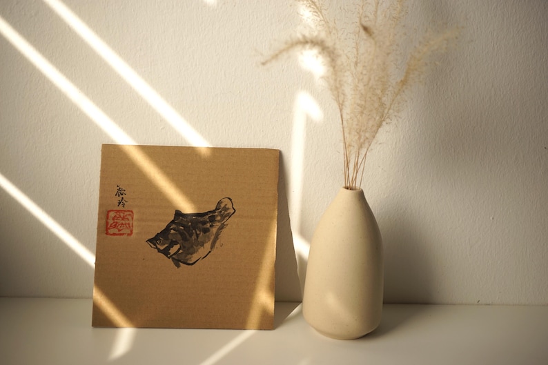May include: A square piece of brown cardboard featuring a black ink wash painting of a fish, accompanied by a red stamp and black calligraphy. Next to it stands a smooth, matte, off-white ceramic vase containing a single sprig of dried pampas grass.