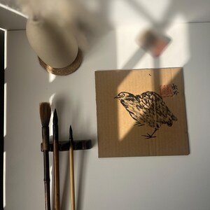 May include: A bird illustration in black and brown ink on a piece of cardboard. The artwork is mounted on a white wall, with calligraphy brushes and a vase visible. The image is lit by natural light.