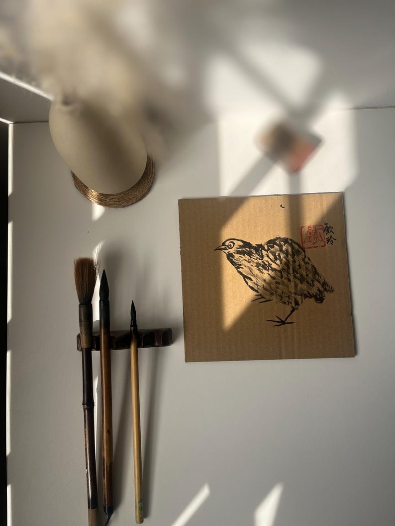 May include: A bird illustration in black and brown ink on a piece of cardboard. The artwork is mounted on a white wall, with calligraphy brushes and a vase visible. The image is lit by natural light.