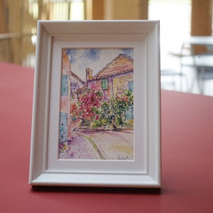 May include: A framed watercolour painting of a European street scene. The artwork depicts buildings with red tile roofs, colourful flowers, and a cobbled street. The frame is white.
