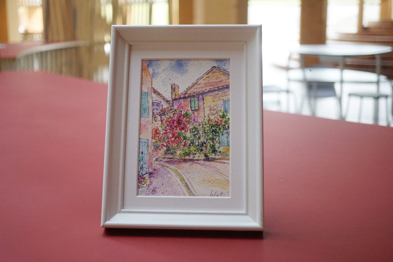 May include: A framed watercolour painting of a European street scene. The artwork depicts buildings with red tile roofs, colourful flowers, and a cobbled street. The frame is white.