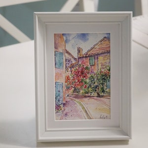 May include: A framed watercolour painting of a European street scene. The artwork depicts buildings with red tile roofs, blue shutters, and vibrant red and green foliage. The painting is in a white frame.
