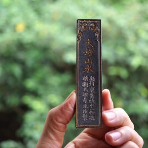 May include: A rectangular, dark-colored object with gold-colored Chinese characters is held in a hand. The object has ornate gold detailing around the edges. The background is a blurred green.