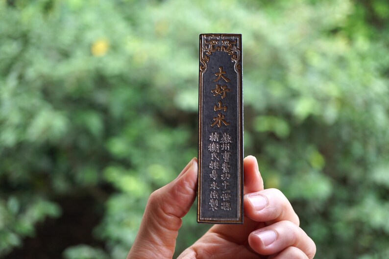 May include: A rectangular, dark-colored object with gold-colored Chinese characters is held in a hand. The object has ornate gold detailing around the edges. The background is a blurred green.