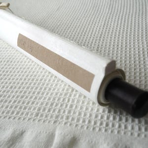 May include: A white rolled scroll featuring a light brown rectangular paper label with faint text. The scroll is secured with a thin beige string tied in a bow at the top and features a smooth black cylindrical end cap on the right side.