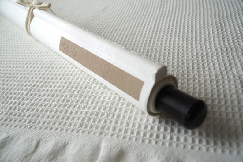May include: A white rolled scroll featuring a light brown rectangular paper label with faint text. The scroll is secured with a thin beige string tied in a bow at the top and features a smooth black cylindrical end cap on the right side.