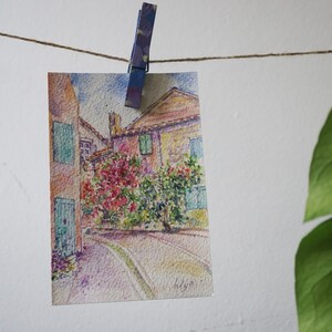 May include: A watercolour painting of a European street scene, clipped to a string with a clothes peg. The artwork depicts buildings with green shutters and red flowers. A green leafy plant is in the background.
