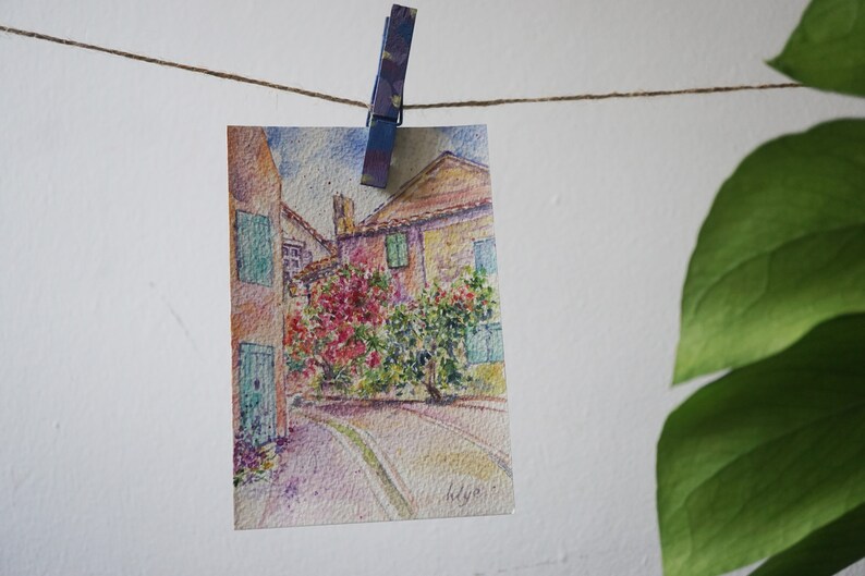 May include: A watercolour painting of a European street scene, clipped to a string with a clothes peg. The artwork depicts buildings with green shutters and red flowers. A green leafy plant is in the background.