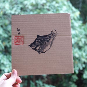 May include: A square piece of brown corrugated cardboard featuring a black ink wash painting of a fish. To the left of the fish, there is black calligraphic text and a square red stamp with an intricate design. The cardboard has a ribbed texture.