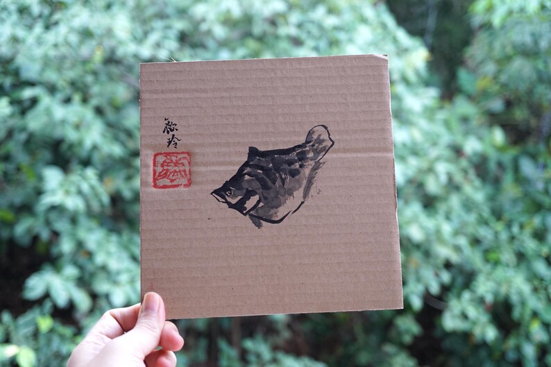 May include: A square piece of brown corrugated cardboard featuring a black ink wash painting of a fish. To the left of the fish, there is black calligraphic text and a square red stamp with an intricate design. The cardboard has a ribbed texture.