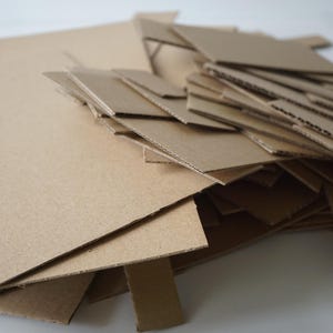 May include: A pile of various-sized, tan-colored cardboard pieces. The pieces are cut into different shapes and sizes, including rectangles and strips. The cardboard is stacked and scattered, suggesting a craft or construction project.