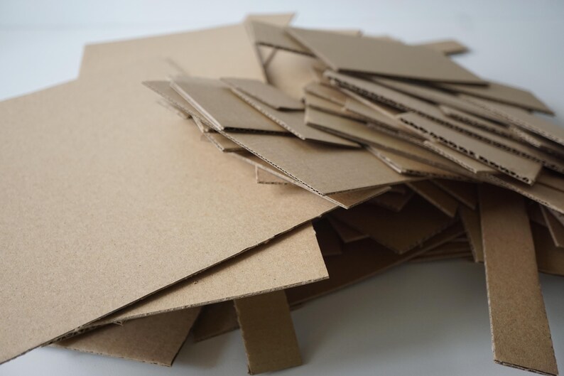 May include: A pile of various-sized, tan-colored cardboard pieces. The pieces are cut into different shapes and sizes, including rectangles and strips. The cardboard is stacked and scattered, suggesting a craft or construction project.