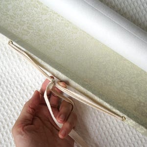 May include: A light green rectangular box with a delicate white floral pattern on the interior lining. The box features a hinged lid secured by a thin, cream-colored fabric ribbon threaded through two small metal eyelets on the side of the container.