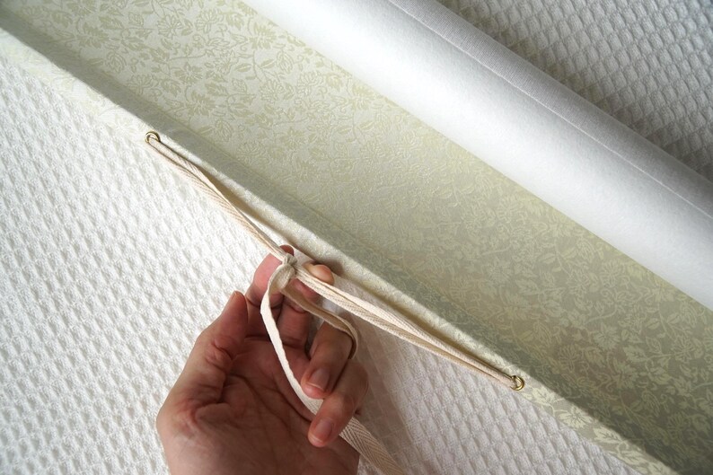 May include: A light green rectangular box with a delicate white floral pattern on the interior lining. The box features a hinged lid secured by a thin, cream-colored fabric ribbon threaded through two small metal eyelets on the side of the container.