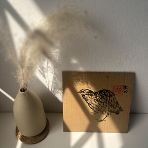 May include: A beige ceramic vase with dried pampas grass and a square cardboard art piece featuring a bird illustration. The bird is depicted in black ink with a red stamp and Japanese characters.