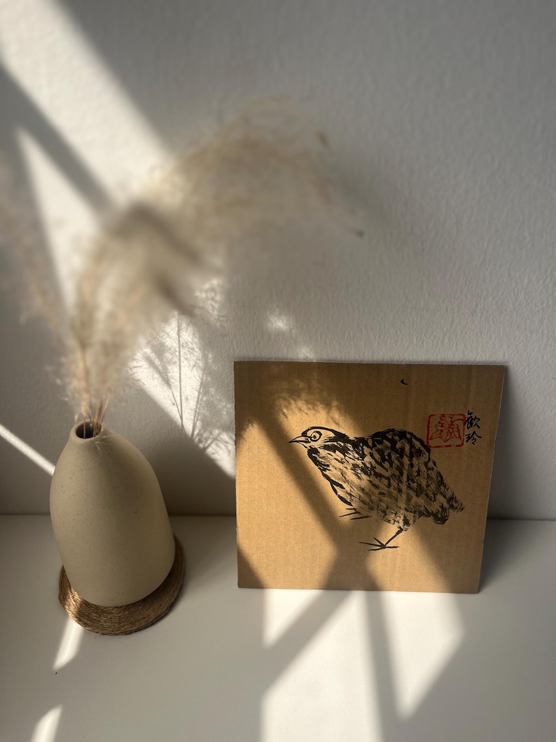 May include: A beige ceramic vase with dried pampas grass and a square cardboard art piece featuring a bird illustration. The bird is depicted in black ink with a red stamp and Japanese characters.