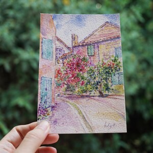 May include: Watercolour painting of a European street scene. The artwork depicts buildings with red-tiled roofs, green shutters, and a cobbled street. Lush pink and green foliage adds colour. The artist's signature is in the lower right corner.
