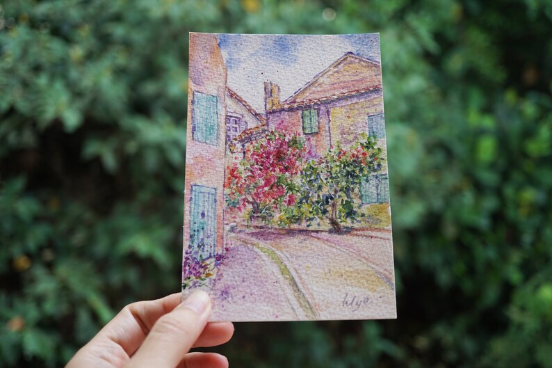 May include: Watercolour painting of a European street scene. The artwork depicts buildings with red-tiled roofs, green shutters, and a cobbled street. Lush pink and green foliage adds colour. The artist's signature is in the lower right corner.
