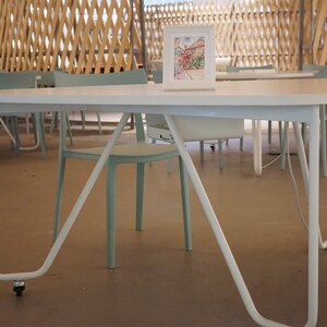 May include: A white oval table with curved legs and a small framed watercolour painting. The table is in a room with light blue chairs and a wooden lattice ceiling. The table has a wheel on one leg.