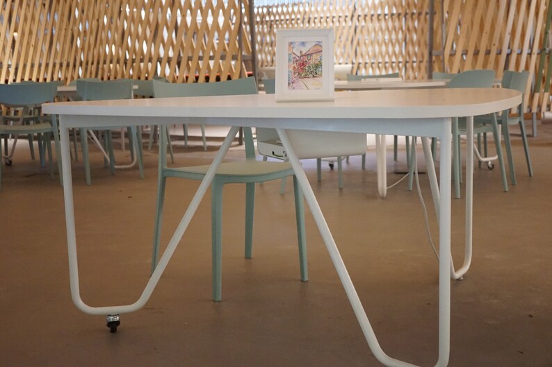 May include: A white oval table with curved legs and a small framed watercolour painting. The table is in a room with light blue chairs and a wooden lattice ceiling. The table has a wheel on one leg.