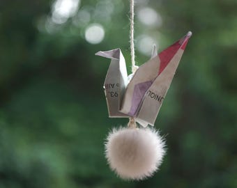 May include: An origami crane folded from printed paper with visible text, featuring a red accent on one wing. The crane is suspended by a thin string and hangs above a small, round, fluffy, light-colored pom-pom ornament.