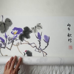 May include: A traditional hanging scroll featuring an ink wash painting of purple flowers and dark grey foliage on a cream-colored paper background. The artwork includes black Chinese calligraphy characters and a small red square seal stamp on the right side.