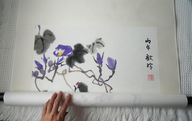 May include: A traditional hanging scroll featuring an ink wash painting of purple flowers and dark grey foliage on a cream-colored paper background. The artwork includes black Chinese calligraphy characters and a small red square seal stamp on the right side.