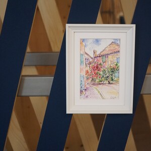 May include: A framed watercolour painting of a European street scene. The artwork depicts buildings with red tile roofs, lush greenery, and a winding road. The painting is in a white frame and is displayed against a blue and wood geometric background.