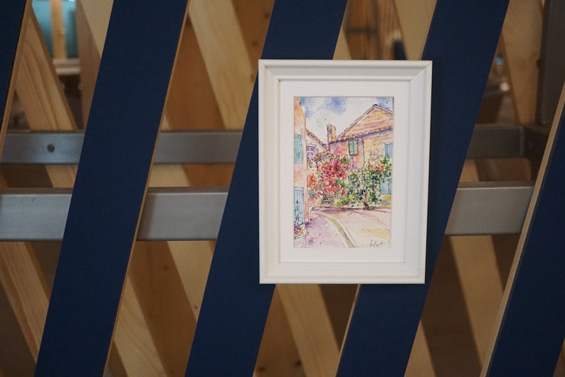 May include: A framed watercolour painting of a European street scene. The artwork depicts buildings with red tile roofs, lush greenery, and a winding road. The painting is in a white frame and is displayed against a blue and wood geometric background.