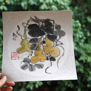 May include: A watercolor painting of gourds and leaves in shades of yellow, green, and black. The artwork features a circular composition with Japanese calligraphy and a red seal. The painting is held in a hand against a backdrop of green foliage.