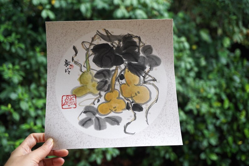 May include: A watercolor painting of gourds and leaves in shades of yellow, green, and black. The artwork features a circular composition with Japanese calligraphy and a red seal. The painting is held in a hand against a backdrop of green foliage.