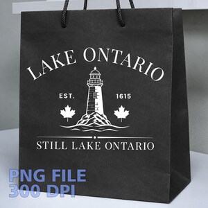 May include: A black paper gift bag with black cord handles featuring a white graphic print. The design includes a lighthouse, two maple leaves, and the text LAKE ONTARIO, EST. 1615, and STILL LAKE ONTARIO. The bag has a matte finish and square base.