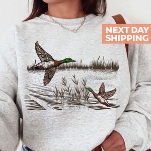 May include: A light gray sweatshirt featuring a detailed illustration of two ducks in flight over a body of water with reeds. The ducks have brown and white wings and green heads. The sweatshirt has a crew neck and long sleeves.