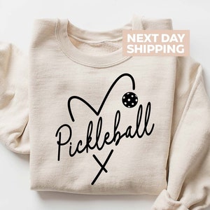 May include: A beige sweatshirt with a black heart design that includes a pickleball and paddles. The text 'Pickleball' is written in a cursive font below the heart. The text 'NEXT DAY SHIPPING' is in a white rectangle in the upper right corner of the image.