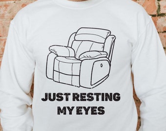 May include: White sweatshirt with a black and white illustration of a recliner and the text "Just resting my eyes".