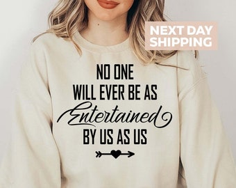 Puede incluir: Una sudadera beige con el texto "No one will ever be as Entertained by us as us" en letras negras. Una flecha con un corazón está debajo del texto.